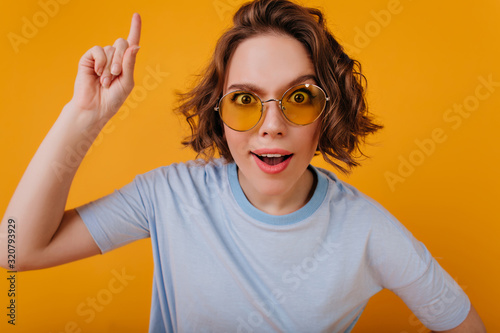 Positive girl wears blue t-shirt expressing funny emotions. Studio photo of short-haired female model in retro sunglasses waving hand with smile.
