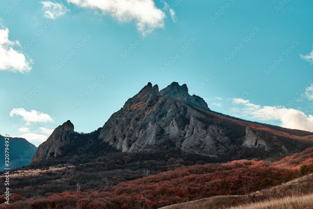 view of a rocky mountain at the foot of which is an autumn forest, the sky is blue with clouds