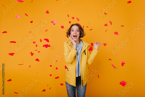 Excited white girl with light-brown wavy hair posing in valentine's day. Indoor photo of happy young woman standing under fallen hearts and laughing.