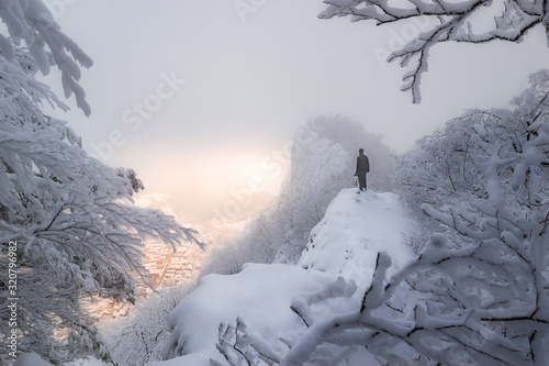 In frosty and foggy weather, girl photographer looks at illuminated picturesque village under snowy mountain