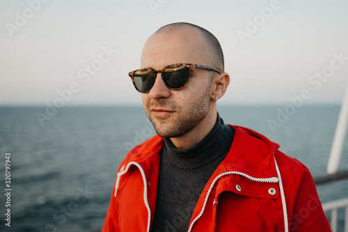 A stylish bald man with stubble in a red jacket in the sunglasses poses aboard a ship looking at the sunset. Close portrait.