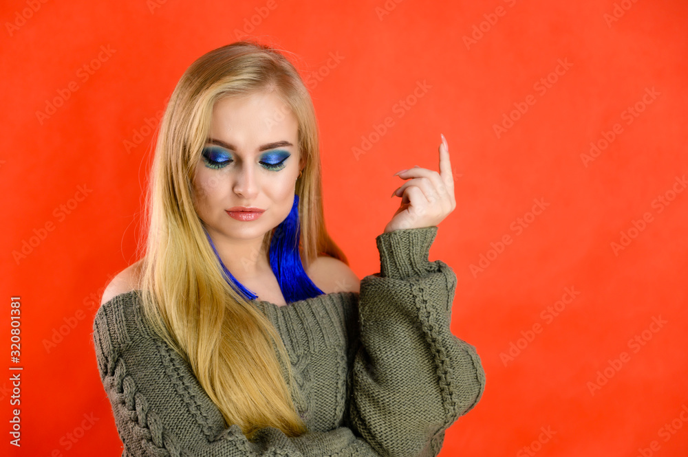 Obraz premium A woman shows different emotions. A pretty blonde model with long hair and excellent make-up is standing in front of the camera on a red background in a green jacket.