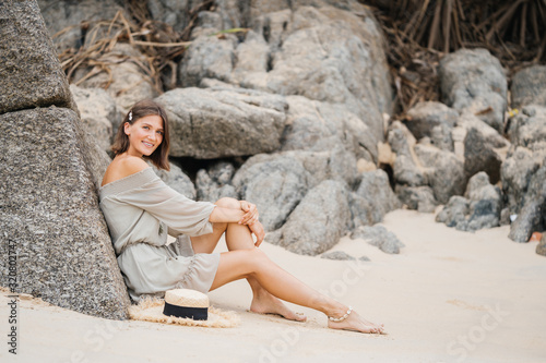 Portrait of a young beautiful girl on the beach near the stone in stylish fashionable clothes with a straw hat sitting on the sand barefoot and enjoying vacation.