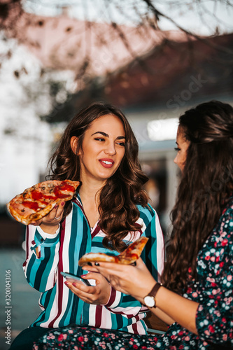 Portrait of two beautiful young women talking and eating pizza outdoors
