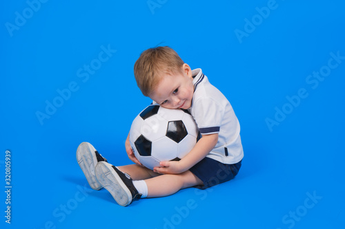 Boy in a football uniform sits on a blue background with a soccer ball and looks forward. © orientka