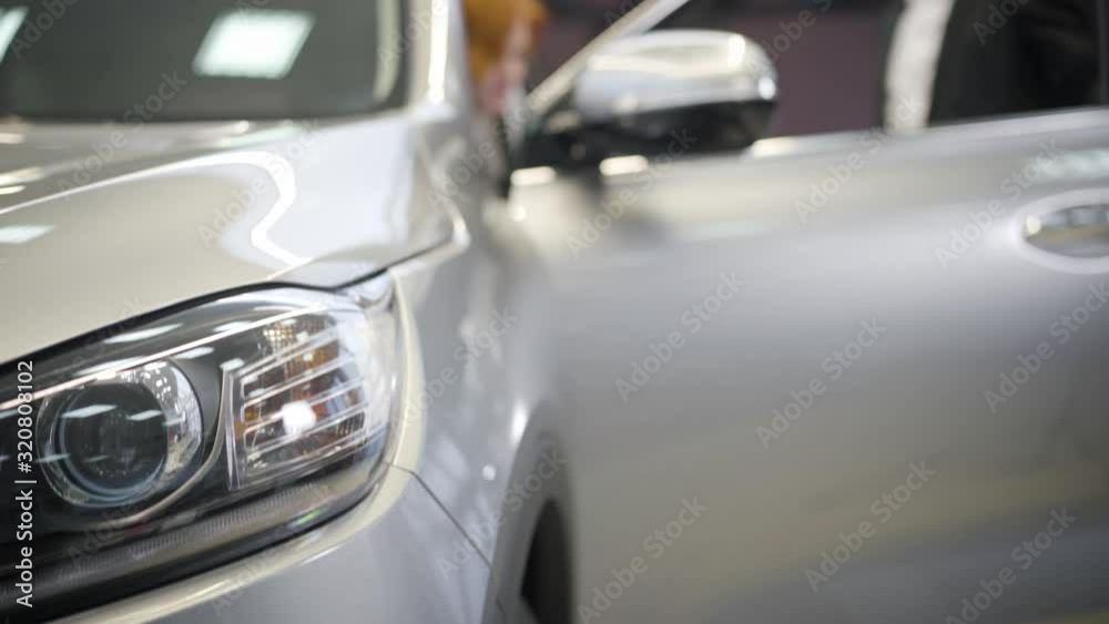 Close-up of car headlamp. Blurred Caucasian man opening door at the background and redhead woman sitting inside. Successful unrecognizable businesswoman buying new automobile. Close-up of car headlamp. Blurred Caucasian man opening door at the background and redhead woman sitting inside. Successful unrecognizable businesswoman buying new automobile.
