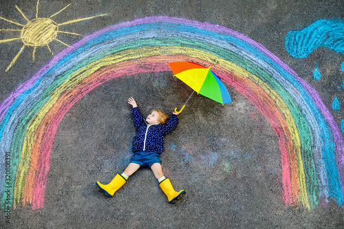 Fototapeta Happy little toddler girl in rubber boots with rainbow sun and clouds with rain painted with colorful chalks on ground or asphalt in summer