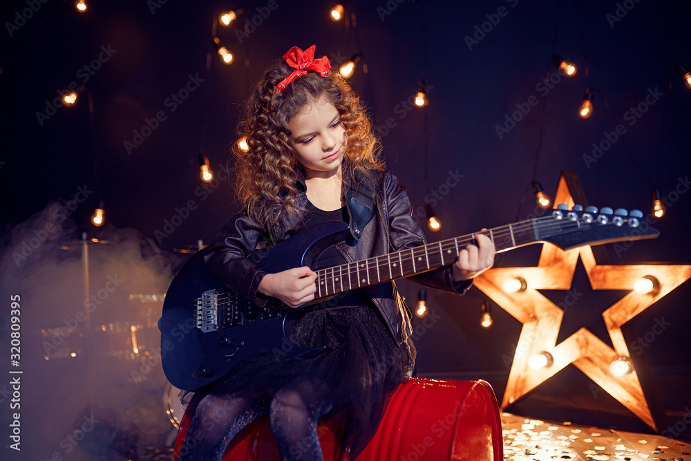 Obraz premium Portrait of a Beautiful rock girl with curly hair wearing leather jacket, boots playing the electric guitar while sitting on a red tank in recording studio.