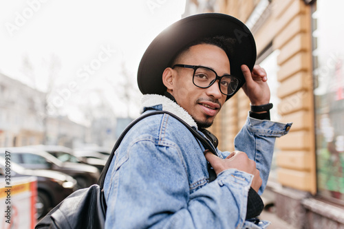 Joyful african male model touching his black hat and looking away on city background. Cheerful man with backpack walking around town in morning.
