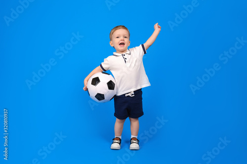 Happy smiling boy stand with soccer ball and rejoices in victory, raising his arms top against blue background. Emotions of winner and champion, place for text. Concept of winning and achieving goal © orientka
