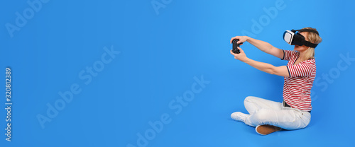 Young girl in white trousers and striped sweater is playing virtual reality with helmet VR and gamepad on a blue background. Innovative technologies and concept of education and entertainment.