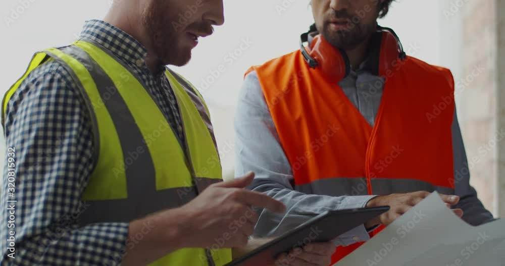 Close up of the two Caucasian men, architect with tablet device and foreman with drawing and plans standing at the building site and talking over the construction. Building concept.