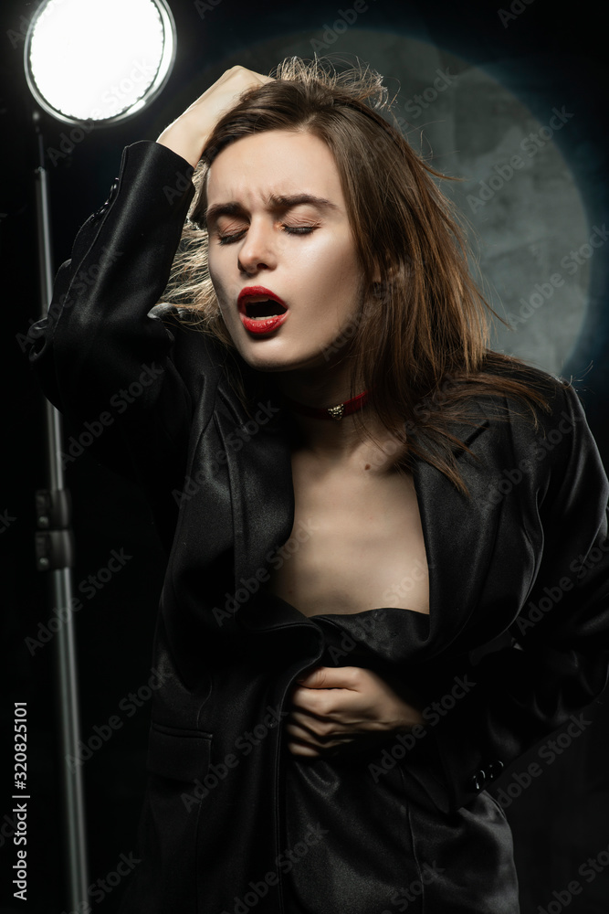 Beautiful slim braless girl, wearing a unbuttoned black blazer, sensually  frowns and touches her hair with her hand on a dark background, posing next  to a light lamp. Advertising, trendy design. Stock