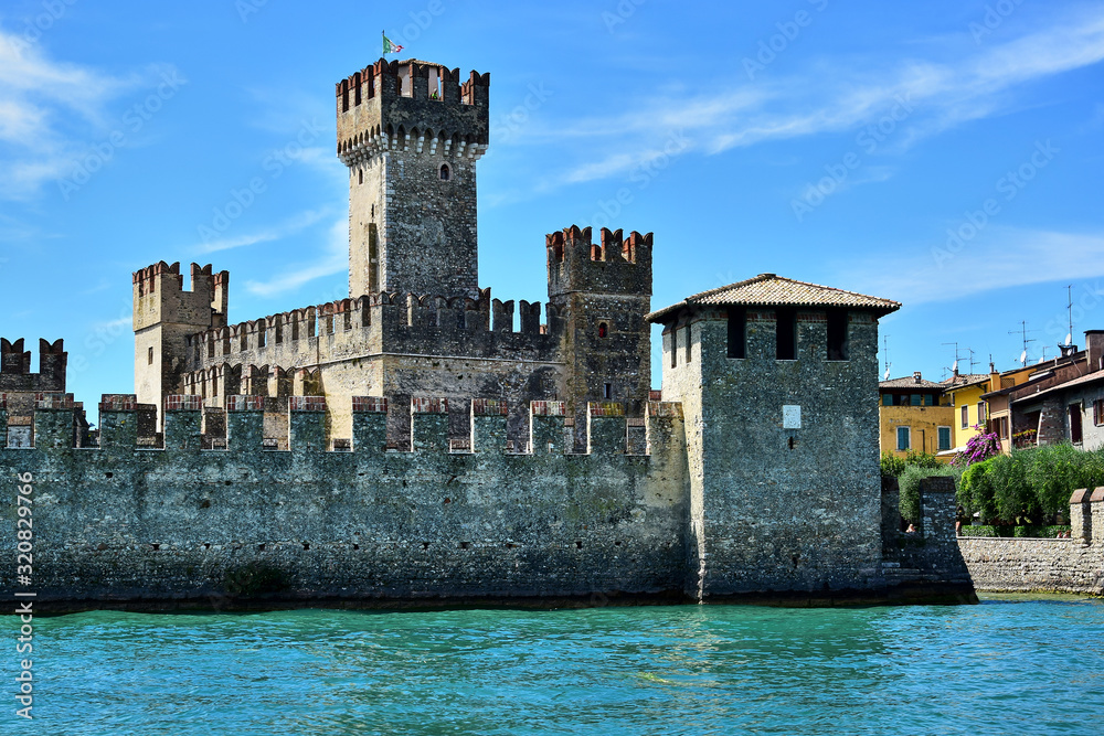 Sirmione, Italy - Scaligero Castle, a fortress on the shores of Lake Garda, stone walls, the Italian flag on the tower, turquoise water and blue sky, in the summer afternoon.