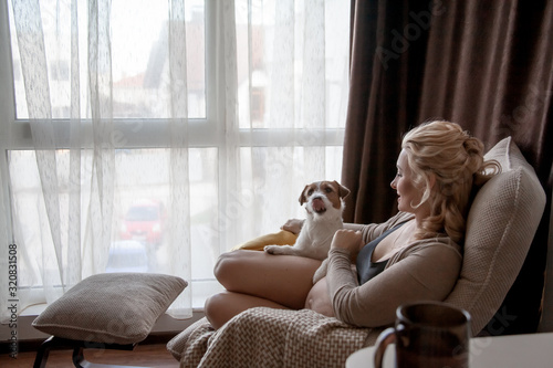 Pregnant woman siting on cozy armchair with cup of tea at home infront of the window. The owner of pet expecting baby and relaxing with dog . Hygge concept.