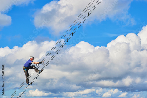 Guy on via ferrata Intersport, Donnerkogel mountains, near Gosau, Austria. Abstract section of a diagonal ladder against white clouds and blue sky - stairway to heaven concept.