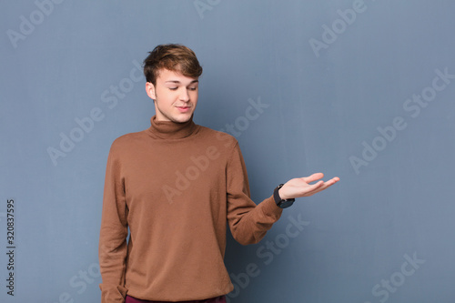 young blonde man feeling happy and smiling casually, looking to an object or concept held on the hand on the side isolated against flat wall photo