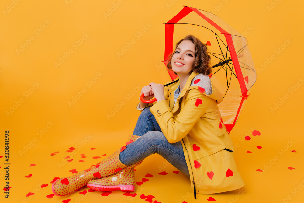 Fototapeta premium Relaxed pretty girl in yellow coat holding paper heart. Indoor photo of wonderful short-haired woman sitting under parasol during photoshoot in valentine's day.
