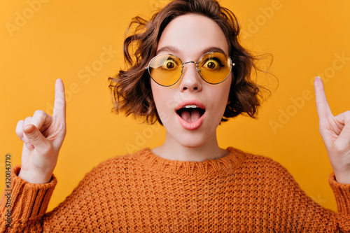 Close-up portrait of positive european woman wears elegant yellow sunglasses. Studio shot of enchanting curly girl expressing surprised emotions.