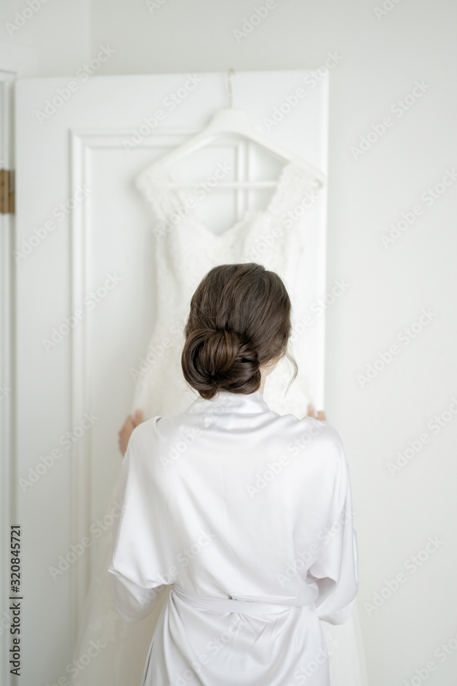 Brunette bride straightens. White dress hanging on a wooden door. Wedding accessories. Brunette bride straightens. White dress hanging on a wooden door. Wedding accessories.