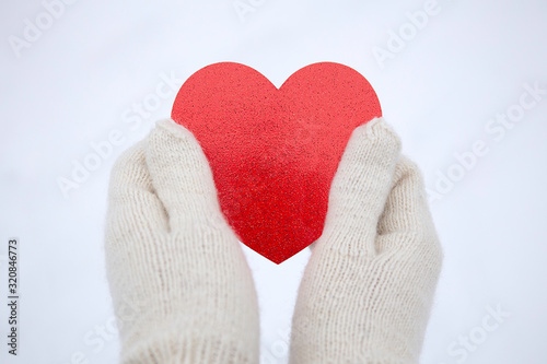 Valentines day background. Hands in white knitted mittens hold a red heart. Red Heart in hands. White snow background. Space for text.