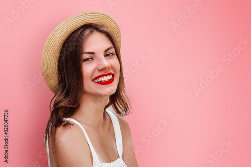 A young girl in straw hat and red lipstick smiling on a pink background, copy space. Tourist girl having fun. Happy girl looking at the camera smiling and laughing, space for text