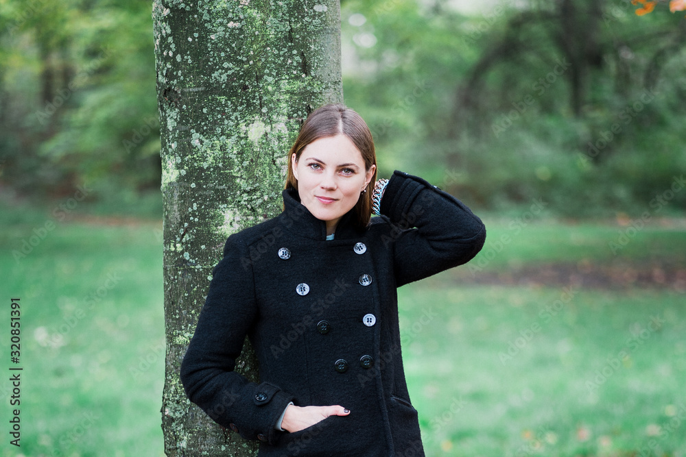 Beautiful woman in a black coat leaning against a tree waiting in the nature, blurred background with copy space Beautiful woman in a black coat leaning against a tree waiting in the nature, blurred background with copy space