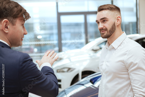 Charming handsome man talking to car dealer while choosing new automobile to buy