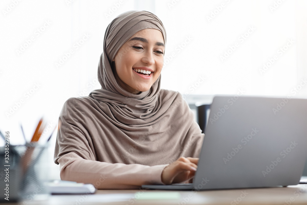 Smiling Arabic Female Entrepreneur In Hijab Using Laptop In Office Smiling Arabic Female Entrepreneur In Hijab Using Laptop In Office