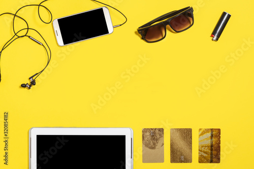 tablet pc, smartphone earphones, cigarette lighter, sunglasses and three gold bank card lying on a yellow background. concept of modern people gadgets and accessories. top view. free copyspace