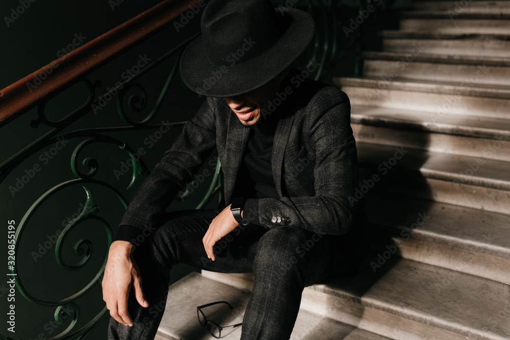 Well-dressed man sitting on the steps with gently smile. Indoor photo of african guy in black jacket and hat. Well-dressed man sitting on the steps with gently smile. Indoor photo of african guy in black jacket and hat.