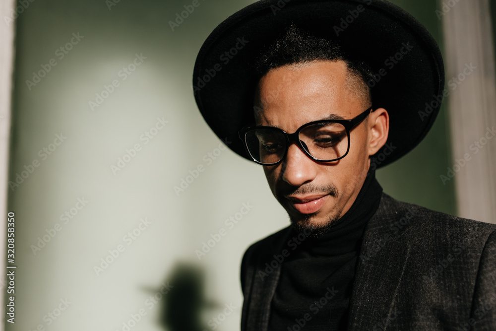 Pensive guy in glasses looking down. Indoor photo of sad african man in hat. Pensive guy in glasses looking down. Indoor photo of sad african man in hat.