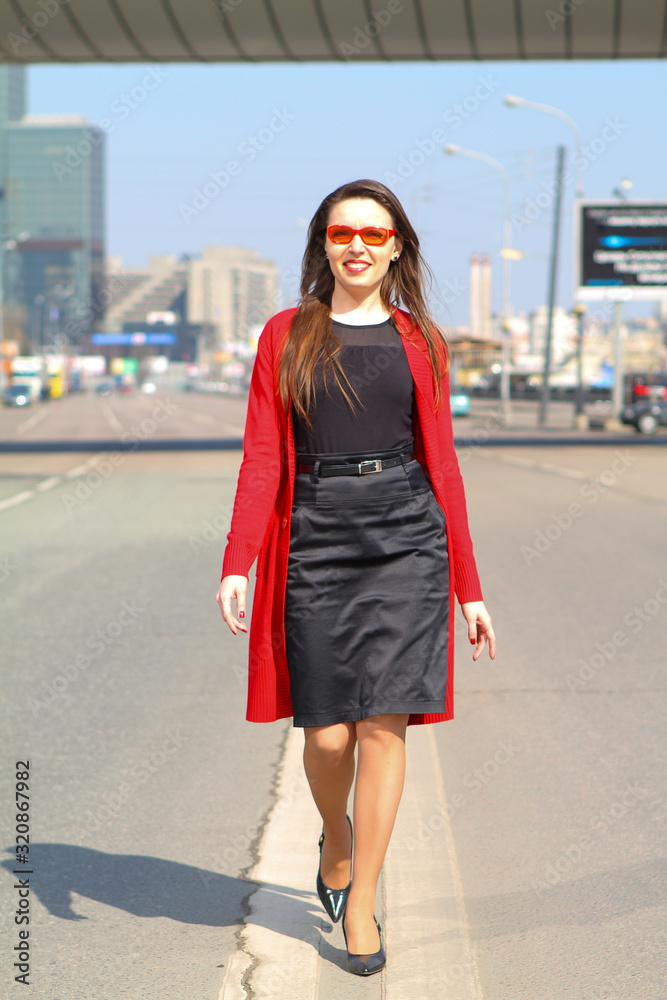 Fototapeta premium A young beautiful happy business lady walking along the dividing line of a city street in clear Sunny weather against the background of office buildings. People, business, beauty.