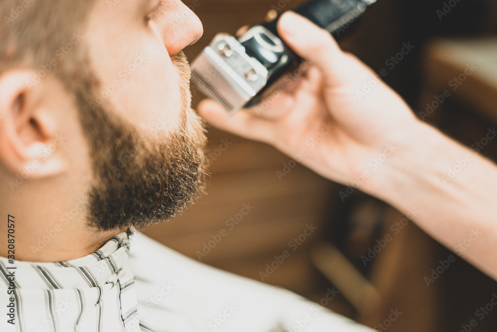 Fototapeta premium Beauty shop for men. Shaving a beard in a barbershop. Barber cuts his beard with a razor and clipper. close up Brutal haircuts. Hairdresser equipment. Selective focus.