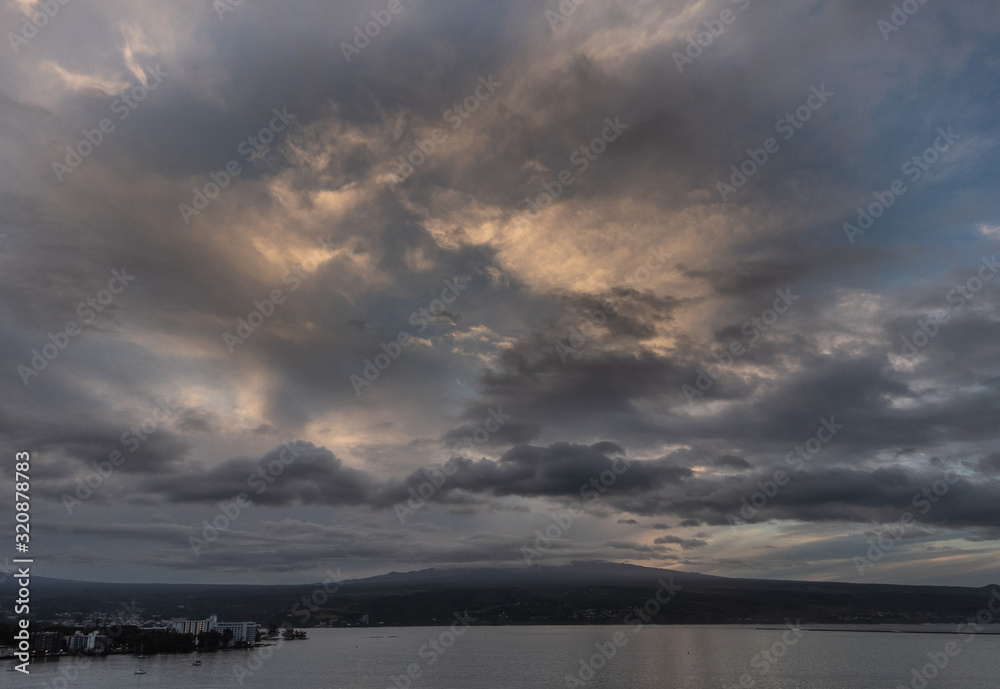 Hilo, Hawaii, USA. - January 14, 2020: Spectacular dark evening cloudscape with yellow sunlight patches over town and volcano on horizon.