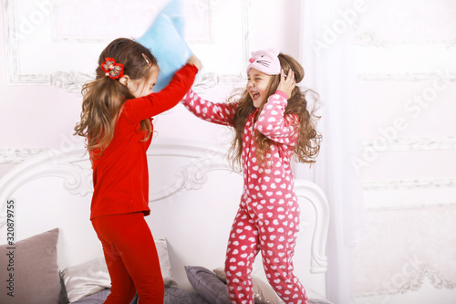 Happy funny children are dressed in bright pajamas playing together and fighting with the pillows on the huge bed