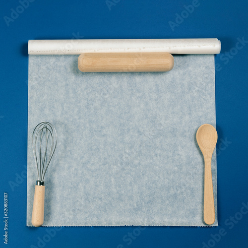 Foto Wooden spoon, rolling pin and balloon whisk over white baking paper on blue background