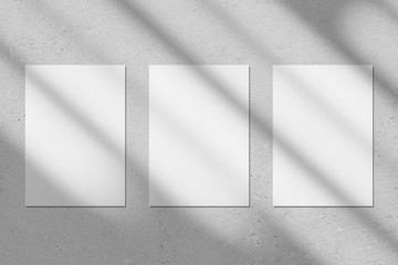 Three empty white vertical rectangle poster or business card mockups with diagonal window shadow on the gray concrete wall.Flat lay, top view. For advertising, brand design, stationery presentation.