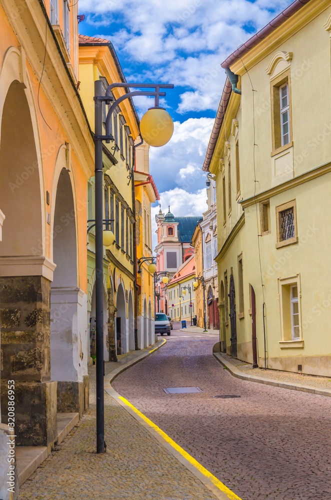 Narrow street in Kutna Hora historical Town Centre with cobblestone road, old style colorful buildings and street lights lamp, blue cloudy sky background, Central Bohemian Region, Czech Republic