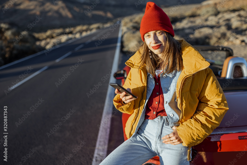 Stylish woman in yellow jacket and red hat enjoying roadtrip, sitting with a smart phone on the car hood on the roadside of the desert valley during a sunset