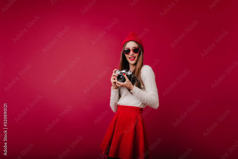 Obraz premium Indoor portrait of interested girl in sunglasses and red hat isolated on claret background. Glamorous female photographer in skirt standing in studio with camera.