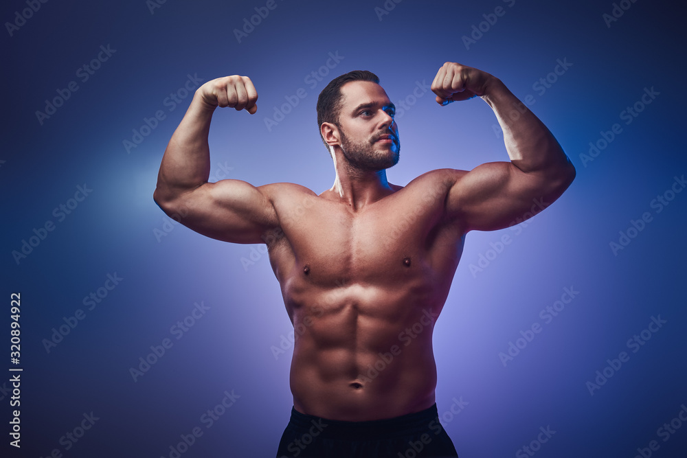 Attractive young bodybuilder is demonstrating his biceps over blue background.