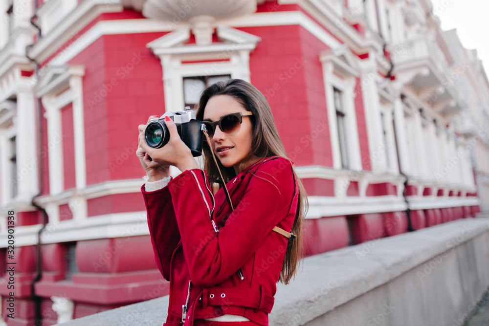 Fototapeta premium Pretty concentrated female photographer spending time outdoor. Beautiful brunette girl in dark sunglasses taking pictures during walk around city.