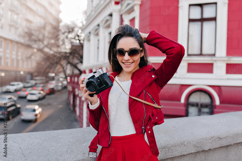 Stunning white woman in sunglasses playing with her hair while posing with camera. Blissful female photographer in red jacket smiling on street background.