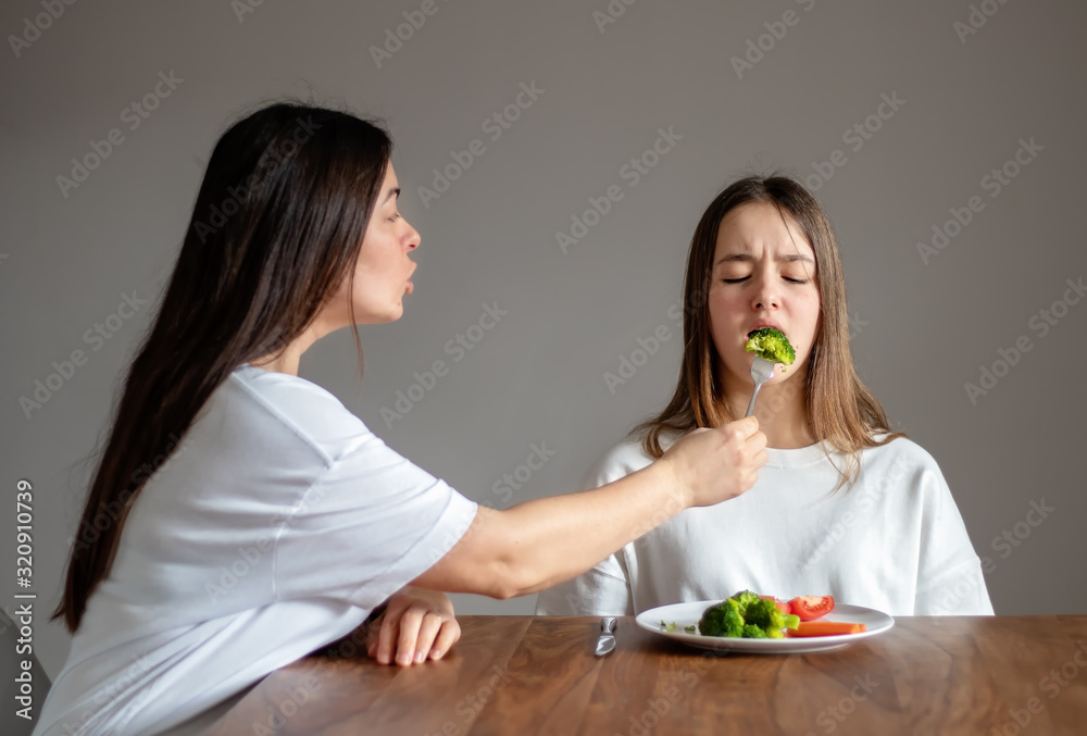Mother forces her teen daughter to eat healthy food feeding her broccoli with fork. Food waste. No vegan diet concept. No choice Mother forces her teen daughter to eat healthy food feeding her broccoli with fork. Food waste. No vegan diet concept. No choice