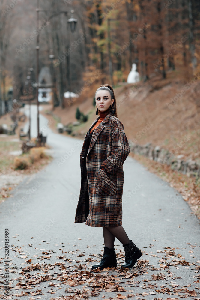 Outdoors portrait of young beautiful fashionable woman strolling in the forest park. Woman beauty autumn fashion. Stylish portrait of a beautiful young woman in a fashion coat in the cage