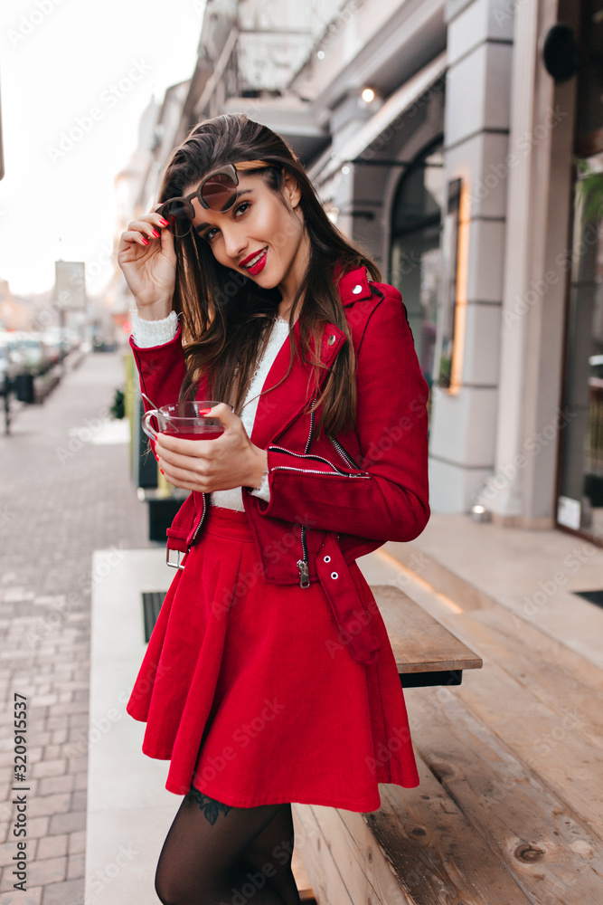 Lovable dark-haired girl in red skirt enjoying outdoor photoshoot. Excited young female model in stylish jacket holding cup of tea on the street.