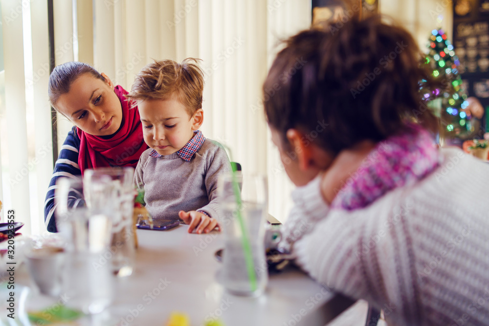 Small little boy caucasian child son sitting in the lap of his mother or aunt while female friend is looking at them teasing smiling holding smart mobile phone playing games making video call family