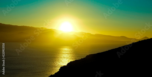 Spectacular sky colors and sunset landscape of a volcanic island with sun reflections in calm ocean.