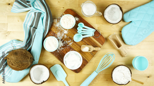 Coconut baking theme flat lay creative layout overhead with coconut flour, milk, oil, and shredded desiccated coconut and baking utensils on modern wooden table.